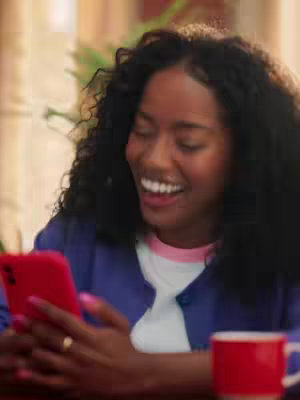 The cover image shows a young woman with curly black hair, smiling and looking at a red smartphone she's holding. She's wearing a white shirt with pink trim and a blue cardigan. A gold ring is visible on her finger. A red mug is on the table near the phone, with some greenery in the background, which suggests she might be indoors. The lighting seems soft and natural.