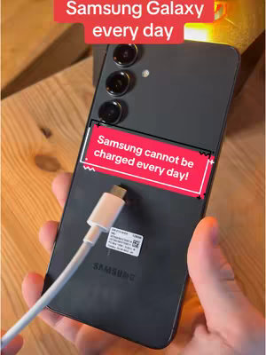 A person is holding a black Samsung Galaxy phone with a white charging cable inserted. The phone's screen faces away from the camera, and the cable’s white connector is plugged into the bottom. Text overlays appear in red boxes: 'Samsung Galaxy every day' at the top and 'Samsung cannot be charged every day!'