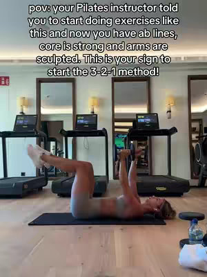 A woman is shown in a gym setting, lying on a black mat and performing an abdominal exercise. Behind her are treadmills and mirrors. Text overlay describes the benefits of the workout, such as having ab lines, core strength, and sculpted arms.