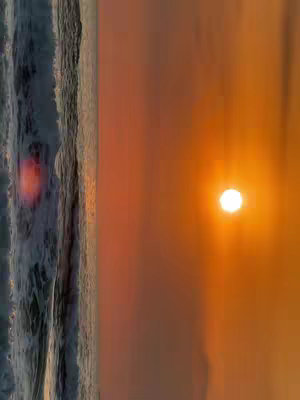 The image shows a seascape at sunset, composed in a vertical orientation. The shot is divided roughly in half, with the left side occupied by the ocean waves meeting the shore, and the right side by a radiant sunset. The water catches the light, with the sky transitioning from orange to lighter hues towards the horizon.