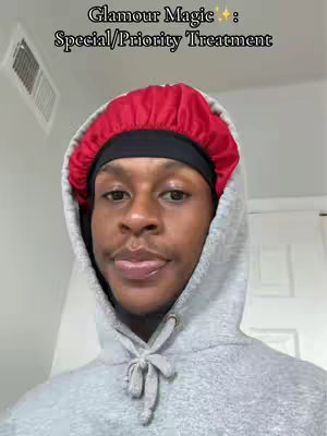 A close-up shot of a Black man wearing a gray hoodie and a red bonnet. He is looking directly at the camera with a neutral expression. The background is plain with a vent. 