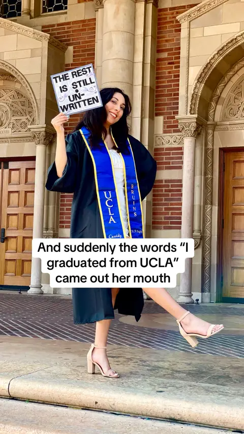 A woman in a graduation gown holds a sign that says, 'THE REST IS STILL UN-WRITTEN' while standing in front of a building.