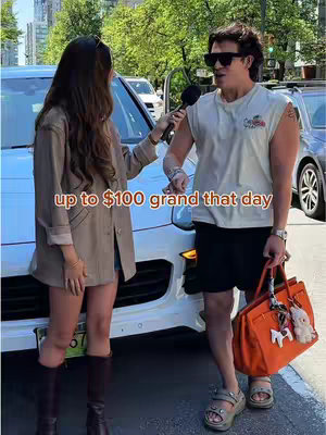 A man and woman stand on a city street near a white car. The man, on the right, wears sunglasses, a white sleeveless shirt, black shorts, and sandals. He holds an orange bag and a mic. The woman on the left wears a blazer, and brown boots, holding a microphone. A text overlay says 'up to $100 grand that day.'