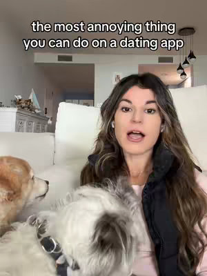 A woman with long brown hair speaks directly to the camera with an open mouth. She is wearing a black vest. In the foreground two dogs are next to her, one is white the other is tan. The background shows a living room setup, with a white couch and a few decorative items.