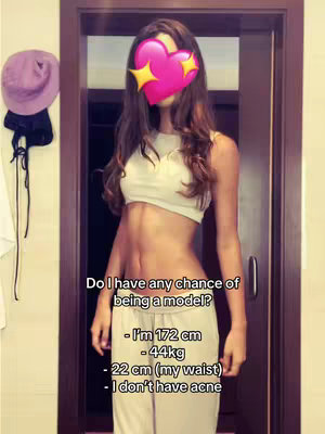 A young woman stands facing the camera, positioned centrally in the frame. Her face is covered by a pink heart emoji overlaid with yellow stars, obscuring her features. She is wearing a light-colored crop top and matching pants. A wooden door frame forms the backdrop behind her, with hints of the room's interior visible. The composition is well-lit, highlighting the subject's silhouette and attire.