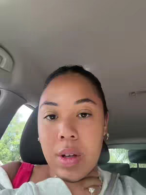 A woman is filmed from the chest up, seated inside a car. The background is an out-of-focus view of a tree line. The woman is facing the camera, with her mouth open, lips slightly parted. She is wearing a light-colored sweater and has an earring. Her hair is pulled back. The interior of the car is visible. The lighting is natural, suggesting daylight.