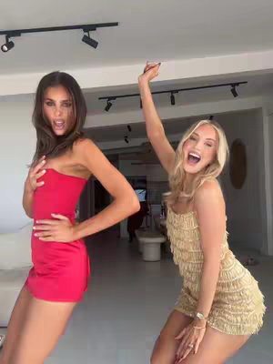 Two young women are captured in a dynamic pose, suggesting movement and energy. The woman on the left is wearing a short, vibrant red dress, holding one hand to her chest. Her dark hair is styled down. The woman on the right is dressed in a fringed gold mini-dress and has her blonde hair styled down. Both women have animated expressions, and the setting appears to be an interior with modern design elements.