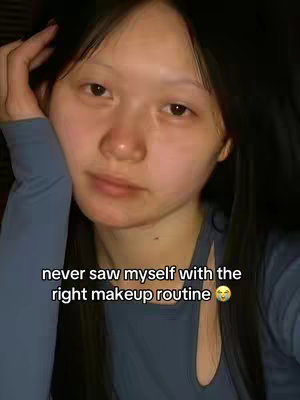 A selfie-style shot featuring a young woman with long, dark hair against a dimly lit background. She is wearing a light blue top and has one hand raised near her head. A text overlay at the bottom reads 'never saw myself with the right makeup routine' in white font.