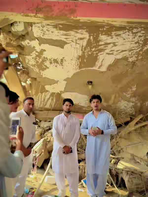 The cover image shows a room that appears to have been partially destroyed. Three men are standing in front of the damaged wall. Two of them are wearing traditional South Asian clothing. The man on the left is holding a phone, possibly recording the scene.