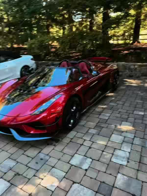 A red Koenigsegg Regera sports car is parked on a cobblestone driveway. The car is the primary focus, presented at a slightly low angle. The background features lush green trees and foliage. There is another white car partially visible behind the main car. The scene is shot during daytime.