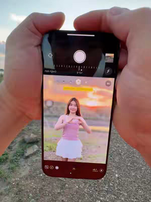 A person holds a smartphone, displaying the camera view focused on a woman posing outdoors. The phone screen shows the woman in a pink top and white skirt against a colorful sunset backdrop. The scene is framed by the hands holding the phone, with a field and sky visible in the background. The phone's camera interface is also visible.