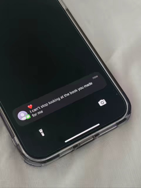 A close-up shot of a black smartphone displaying a message conversation. The phone is in a clear case, and the background appears to be a white fabric. The message bubble reads 'I can't stop looking at the book you made for me,' accompanied by a heart emoji and a profile picture icon. The time 'now' is displayed in the top right corner.