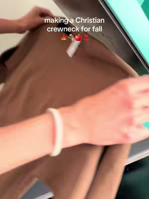 The cover image shows a person's hands holding a brown crewneck sweatshirt. Overlay text at the top reads, 'making a Christian crewneck for fall,' with small emoji graphics. The setting seems to be indoors, possibly a workspace.