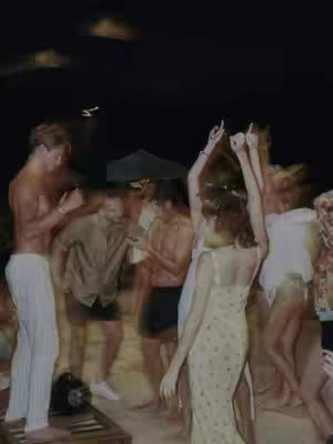 A blurry, nighttime photo shows a group of people dancing and interacting at what appears to be an outdoor party or gathering. In the foreground, a person in an off-white dress faces away from the camera with their arms raised. Other individuals, mostly male, are visible in various poses dancing or standing in the surrounding area. The lighting is dim