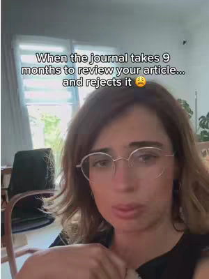 The cover image shows a woman with brown hair and glasses, looking somber. Text overlayed on the image reads, "When the journal takes 9 months to review your article... and rejects it 😩". The background includes an out-of-focus window, chair, and plant.
