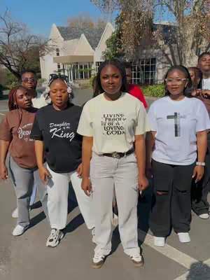 A group of young women stand outdoors on a sunny day, posing for the camera. The background features a large white building with a gabled roof and several trees. The women are dressed in casual attire, including t-shirts with various religious and inspirational phrases screen-printed on them, along with jeans and sneakers. The composition is a medium shot, capturing the group from the torso up, with natural lighting.
