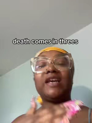 A woman with clear framed glasses and a yellow head covering, looks directly at the camera, which is angled upwards. The text overlay 'death comes in threes' appears in the upper portion of the frame. She is in front of a plain wall with some of her upper body present.