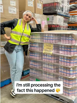 A woman is standing in what appears to be a warehouse setting. She poses next to a stacked pallet of pink-labeled products, possibly for a beauty brand. She is wearing a yellow or neon vest over a black jacket and blue jeans, with her hair neatly contained under a white hairnet. The background includes boxes and other storage items, indicating an industrial setting. Below the woman, overlay text reads, 'I'm still processing the fact this happened 😲'.