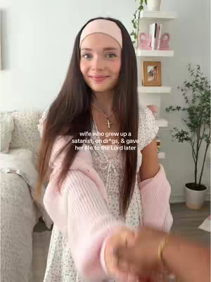 A young woman with long brown hair and a pink headband is smiling at the camera. She is wearing a white floral dress and a pink cardigan. Her right hand is being held by another person who is just out of frame.