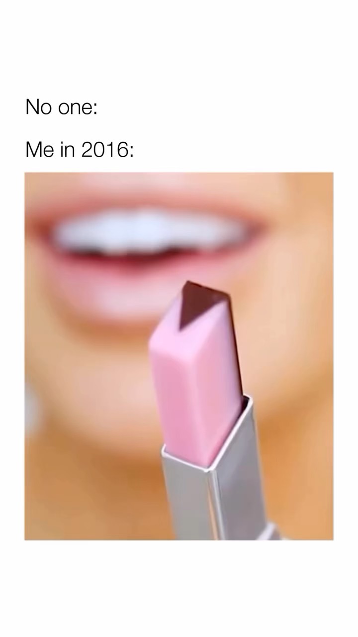 A close-up photo with a white background. In the foreground, a lipstick with a pink base and a brown triangle tip is held. Slightly blurred behind the lipstick is a close-up of a person's mouth, with visible white teeth and a small part of the upper lip, creating a shallow depth of field.