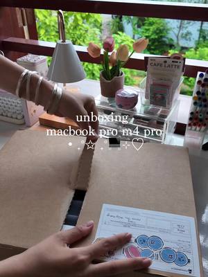 A brightly lit scene from an unboxing video, primarily showing two hands interacting with a cardboard box. The hands, adorned with multiple silver bracelets on the left wrist, are in the process of opening the box, revealing a label with colorful stickers underneath. The background features elements of a workspace, glimpses of foliage and a white stylish lamp.