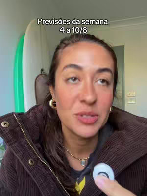 A woman with brown hair and a brown jacket is speaking in front of a background with a white surfboard and green wall.