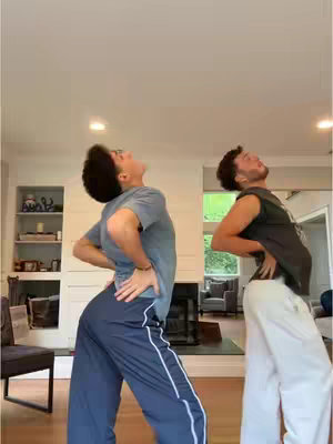 Two young men are shown in a domestic interior, possibly a living room. They are leaning backward with their hands on their lower backs, as if posing or stretching in a dance-like gesture. The background shows a white wall, a fireplace, and a touch of a backyard, suggesting a casual or home setting.