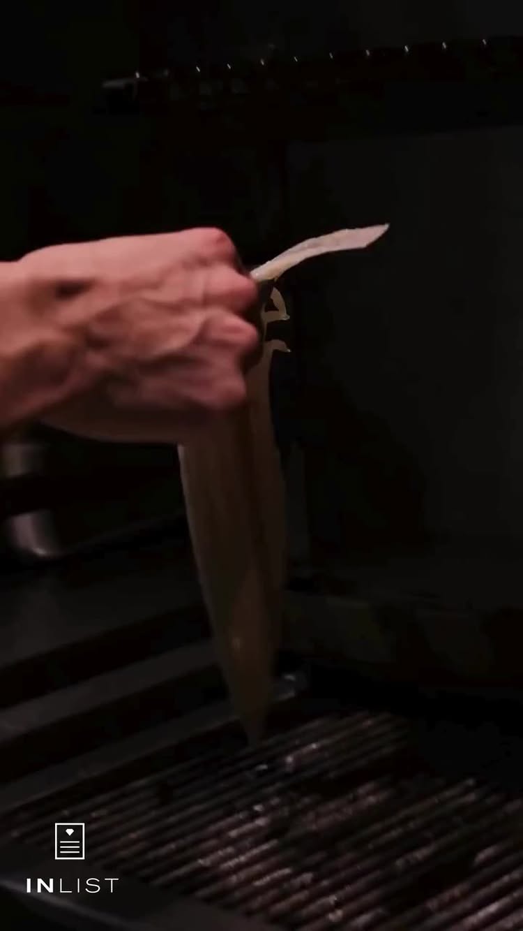 The cover photo is a close-up shot of a person's hand holding a piece of food over a grill. The hand is positioned in the upper left, holding a long, thin, off-white piece of food, possibly a type of seafood. Below the food, a dark grill with horizontal bars is visible, suggesting the food is being prepared. The background is primarily dark, with subtle lighting and shadows that emphasize the grill and the hand. The InList app logo is placed on the bottom left.