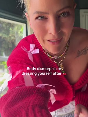 A selfie-style shot featuring a woman with short, blonde hair looking towards the camera. She's wearing a red cardigan with bows and several necklaces. The background is a blurred view of a window and greenery. Text overlayed reads 'Body dysmorphia or dressing yourself after'. The composition focuses on the woman's face and upper body.