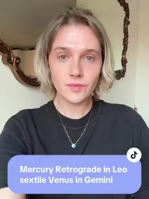 A person with light skin, short blonde hair, and light eyes gazes directly at the camera. They wear a black top and a silver necklace with a blue pendant. A light purple banner at the bottom displays white text related to astrology. In the blurred background, there's a mirror and decorative elements.