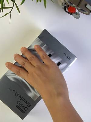 A top-down shot showcases a person's hand gently resting on a silver OPPO phone box. The box is angled, partially obscuring the white surface beneath. A vibrant green plant and a small gray toy are visible in the background.