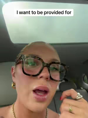 The cover image shows a close-up of a person’s face inside a vehicle, likely a car. Overlayed text reads 'I want to be provided for'. The person is looking at the camera with a slightly open mouth. They have large, square-framed glasses, are wearing earrings and rings, and have a necklace. The lighting appears to be natural, shining in from the vehicle’s sunroof.