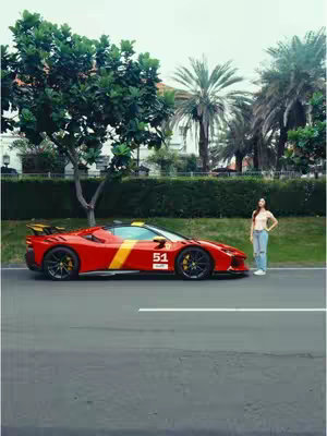 An eye-level shot shows a red sports car, parked on an asphalt road with a white line. A woman stands next to the car, with a green leafy background.
