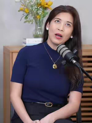 A woman sits in front of a microphone, facing the camera. She wears a navy blue top and her dark hair is styled around her face. A floral arrangement in a vase sits behind her.