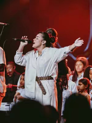 The cover image shows a woman with dark hair holding a microphone and singing on stage with both arms raised. She is wearing a white outfit and a belt with decorative circles. Other people are in the blurred background, illuminated by warm stage lighting. The overall composition suggests a live worship moment.