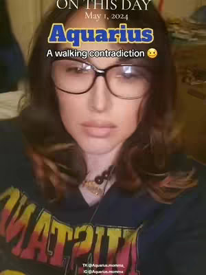 A selfie-style shot featuring a woman with brown hair and glasses. Text overlays include the date 'May 1, 2024,' and astrology-related phrases: 'Aquarius' and 'A walking contradiction'. The woman is facing the camera with a neutral expression, wearing a dark t-shirt. The background is slightly blurred.