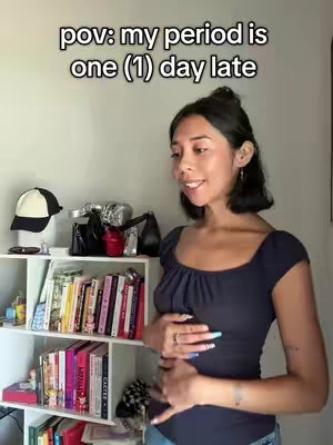 A woman, with dark hair and a navy blue top, stands smiling in front of a shelf with books and decorations. The text overlay reads, 'pov: my period is one (1) day late'. She's gently touching her lower stomach.