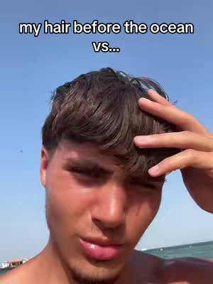 A young man with short, dark hair is looking at the camera, his skin slightly flushed as if he's been in the sun. He is outdoors with a blue sky and water visible behind him.