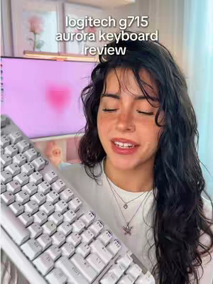 A young woman holds a white Logitech G715 Aurora keyboard, looking at the camera with her eyes closed and a slight smile. The keyboard is in the foreground, taking up a significant portion of the frame. Behind her, a computer monitor displays a blurred pink image, possibly a heart. The background suggests a brightly lit room with a casual, intimate setup, with decorative elements visible towards the top left of the frame.