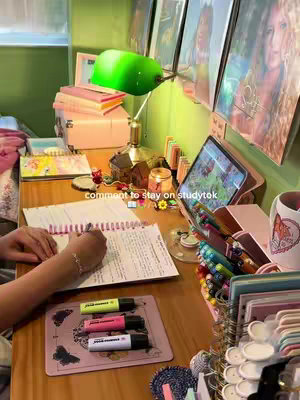 A desk with books, iPad, stationery, and decorative items. A person's hand is writing in a notebook. The scene is lit by a green desk lamp with a candle and framed pictures in the background.