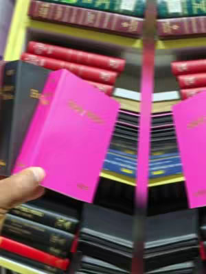 The image shows a hand holding a bright pink book with gold lettering. There are other colorfully bound books, including black and red ones and books in stacks, indicating a bookstore setting.