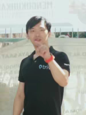 A man in a black polo shirt with a logo is gesturing with his hand raised, possibly pointing or indicating something. The background appears to be a light-colored, possibly outdoor, setting.