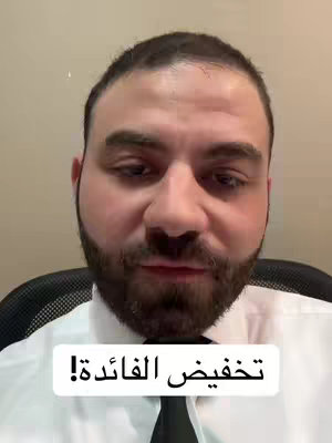 A man with a beard is in the foreground in a button-down shirt and tie facing the camera. The background is a light tan wall. There is a white rectangle at the bottom of the screen with Arabic text.