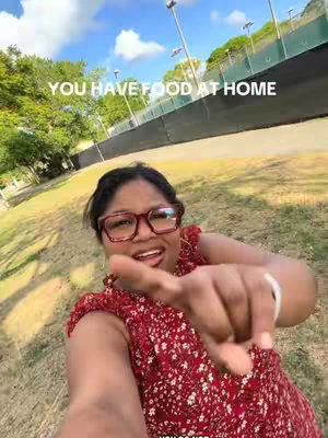 A woman with dark hair and glasses is pointing towards the viewer with a finger. She is outdoors, in a green space, with trees and a blue sky visible in the background. Text overlays the image.