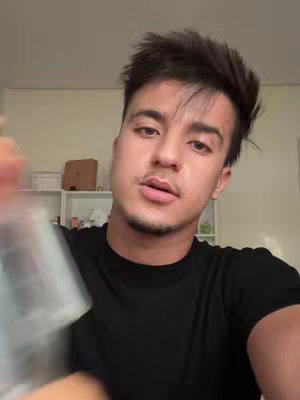 A selfie-style shot of a young man looking directly at the camera. He is in a room with a white wall and some shelves are visible in the background. He is wearing a black t-shirt. A clear bottle is held in the foreground, partially obscuring the shot.