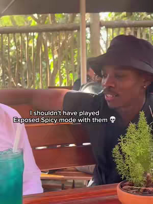 A man in a black hat and shirt is seated outside, gesturing with his left hand while talking. Text overlay reads, "I shouldn't have played Exposed Spicy mode with them". A small plant in an orange pot and a blue drink in a glass are on the table.