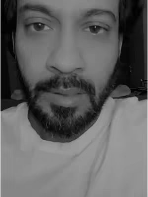 A close-up black and white portrait of a man looking directly at the camera. He has dark hair, a full beard, and is wearing a white t-shirt. The lighting is soft, and the focus is on his face.