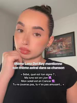 A selfie-style shot of a young woman with light makeup on. She is looking at the camera with slightly pursed lips. The background includes a wall with other framed art, and the woman is wearing a dark top. Text overlay includes a quote from Lana Del Rey.