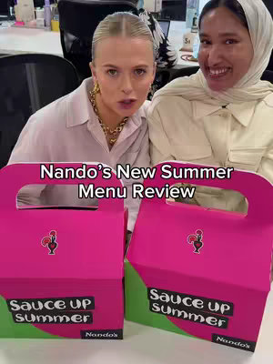 Two women are seated, facing the camera. They are in front of boxes of food. Overlayed on the image is the text "Nando's New Summer Menu Review". The women are smiling towards the camera.