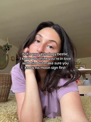 A selfie-style image featuring a woman inside a cozy-looking room. She's positioned in the foreground, slightly off-center, with her face prominently displayed. The woman has dark hair and is wearing a light purple top. She's looking at the camera with a relaxed expression. The background is blurred, but we can see elements like a neutral-colored wall, a portion of the ceiling, and what appears to be houseplants. A text overlay towards the mid-top emphasizes the astrology theme.